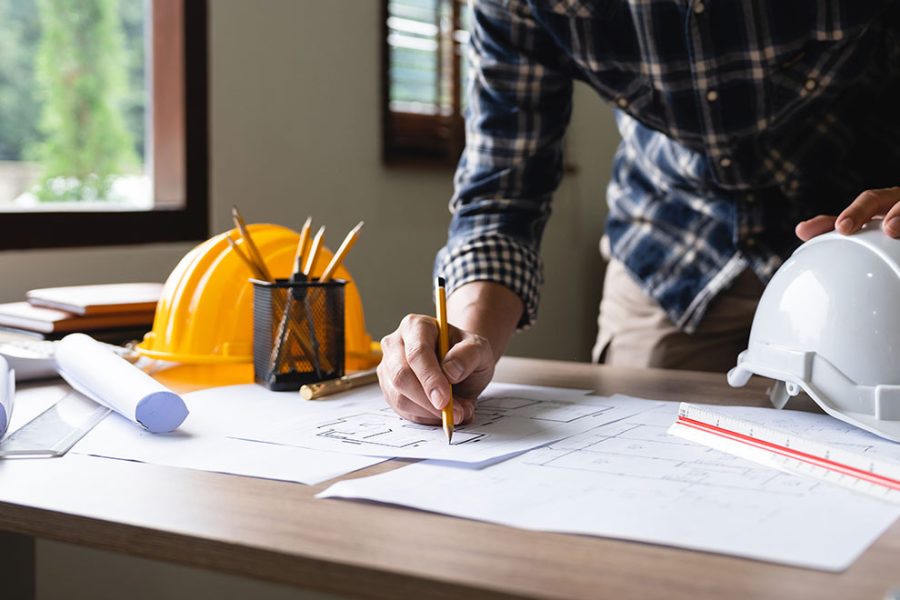 um engenheiro anotando algumas informações na planta de um projeto residencial e um capacete junto de um escalímetro ao lado, com vários papéis ao redor da mesa de madeira