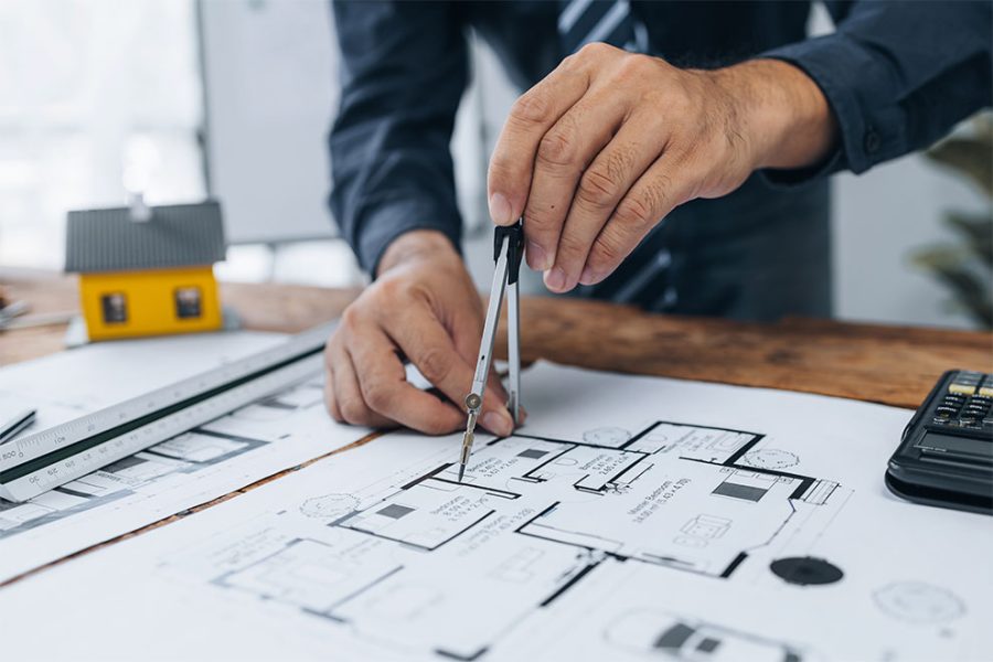 um engenheiro usando um compasso em cima de um projeto residencial com um escalímetro e uma calculadora ao seu lado em cima da mesa
