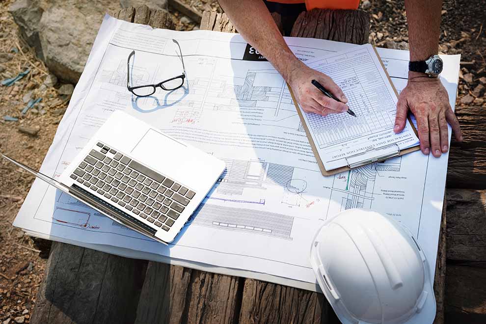 um engenheiro anotando em uma prancheta alguns dados sobre o projeto, em baixo o memorial descritivo do projeto com o capacete, um óculos e um notebook em cima de uma tábua de madeira